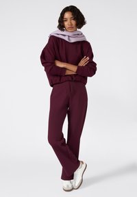 Bordowy sweatshirt z opadającymi ramionami, zestawiony z dopasowanymi spodniami o zwężającym się kroju. Jasnoliliowy szalik opada na szyję. Białe sneakersy.