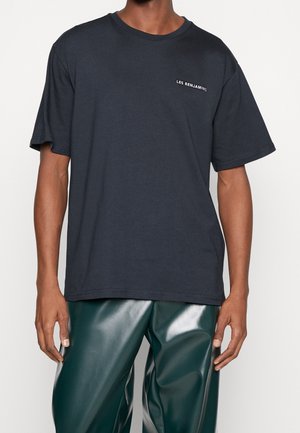 Mężczyzna ubrany w ciemnogranatowy, oversize’owy T-shirt z napisem „LES BENJAMINS” oraz błyszczące ciemnozielone skórzane spodnie, na jednolitym tle.