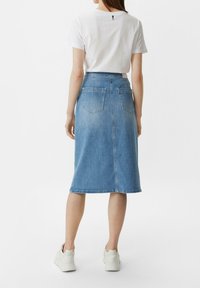 Kobieta w białych trampkach, białej koszulce z krótkim rękawem i średniej długości niebieskiej spódnicy dżinsowej, stojąca tyłem na białym tle.