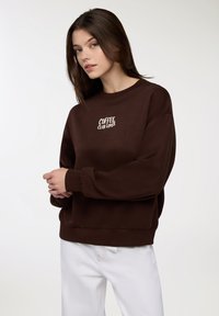 Młoda kobieta z brązowymi włosami nosi ciemnobrązową bluzę z napisem "COFFEE CLUB LOVER" i białe spodnie na tle jasnojednolitego tła.