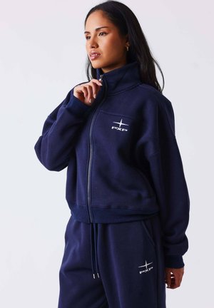 Femme aux longs cheveux foncés portant une veste zippée bleu marine et un pantalon assorti avec des logos blancs PXP, debout devant un fond clair uni.