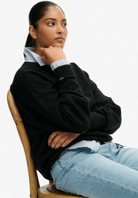 Zwarte chunky gebreide trui over een lichtblauw gestreept blouse met kraag, gecombineerd met lichtblauwe jeans, zittend op een houten stoel.