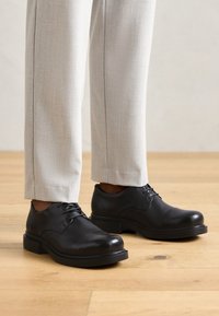 Fekete bőrdresszcipők kerek orral, fűzős kivitelben, vastag fekete gumitalppal, világosszürke testre szabott nadrággal párosítva.