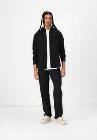 Svart skjorte med knapper, hvit t-skjorte under, svarte bukser, lyse sneakers. Modellen står foran en ensfarget hvit bakgrunn.
