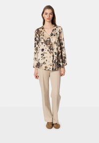 Blusa stampata con scollatura a V e maniche lunghe in beige, caratterizzata da macchie di leopardato nere e marroni. Abbinata a pantaloni beige chiaro e scarpe marroni.