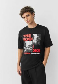 Camiseta de algodón negra con un diseño gráfico que incluye texto e imágenes en rojo y blanco, con elementos musicales y tipografía en negrita.