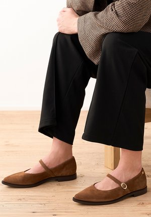 Osoba siedząca, ubrana w brązowe zamszowe baleriny Mary Jane z paskiem na sprzączkę, czarne spodnie i brązową kurtkę we wzory.