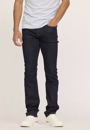 Jean en denim bleu foncé avec une coupe slim et une jambe droite. Présentant des coutures orange et des poches avant, associés à des baskets blanches.