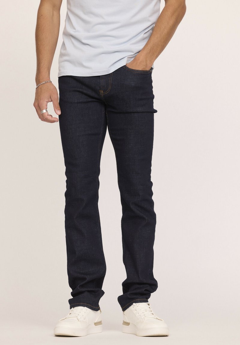 Jean en denim bleu foncé avec une coupe slim et une jambe droite. Présentant des coutures orange et des poches avant, associés à des baskets blanches.