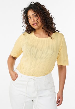 Lockige Frau, die ein gelbes kurzärmeliges Strickoberteil und weiße, hoch taillierte Hose trägt, mit den Händen in den Taschen.