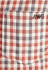 Tissu à carreaux rouge, blanc et gris avec une poche poitrine arborant un logo brodé noir "A J" et un emblème d'aigle.