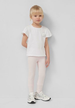 Young girl with blonde hair wearing a white short-sleeve shirt, light pink leggings, and white sneakers standing against a plain gray wall.