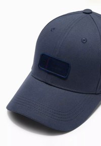 Granatowa czapka wykonana z materiału, z haftowaną metką z przodu, zakrzywionym daszkiem i sześcioma otworami wentylacyjnymi. Gładka tekstura.