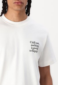 Weißes Baumwoll-T-Shirt mit einem schwarzen Textdruck auf der linken Seite, der "Entspanne dich, nichts wird passieren" lautet.