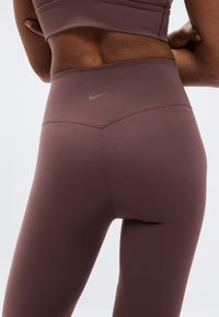 Κολάν Nike σε πλούσιο μωβ χρώμα, με ψηλή μέση και λεία υφή. Διακριτική λεπτομέρεια λογότυπου πάνω από τη μέση.