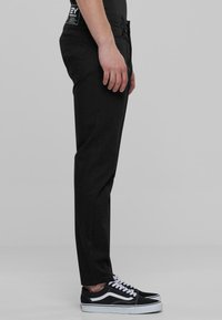 Musta slim-fit-housut sileällä pinnalla, vetoketjukiinnitys ja sivutaskut, yhdistettynä mustavalkoisiin tennareihin. Sivuprofiili.