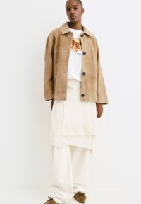 Tan suede jack met knopen, wit graphic t-shirt, crème gelaagd rokje en bruine instappers. Geweven stof en losse pasvorm.