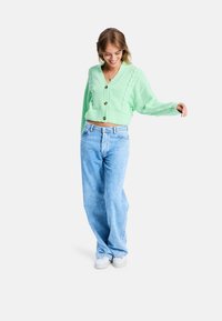 Mintgrüner, gestrickter, kurz geschnittener Cardigan mit V-Ausschnitt, der über Kabelstrickmuster und braune Knöpfe verfügt, kombiniert mit hellblauen, weit geschnittenen Jeans.