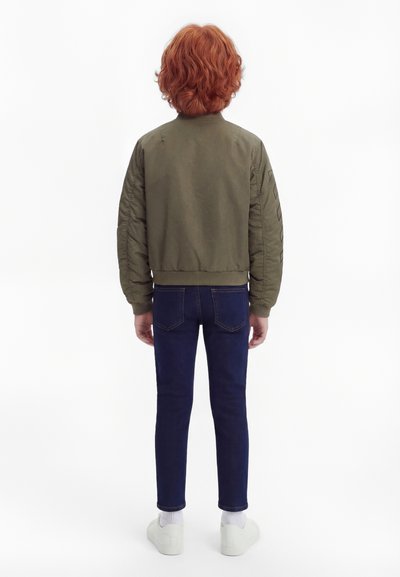 Παιδί με κόκκινα σγουρά μαλλιά στέκεται γυρισμένο προς τα πίσω, φορώντας ένα μπουφάν μπόμερ ελιάς, σκούρα μπλε τζιν, λευκές κάλτσες και λευκά αθλητικά παπούτσια.