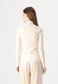 Maglietta lunga beige chiaro con colletto arricciato, texture liscia e cucitura centrale sul retro, caratterizzata da un piccolo dettaglio nero al collo.