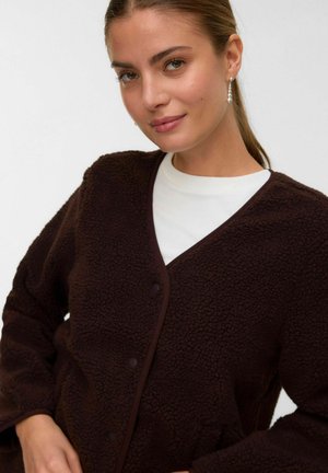 Jeune femme portant des boucles d'oreilles en perles, un haut blanc et une veste en polaire texturée marron foncé avec des boutons pression.