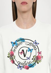 Femme portant un sweat-shirt blanc avec un motif floral et papillons circulaire entourant le texte "Versace Jeans Couture."