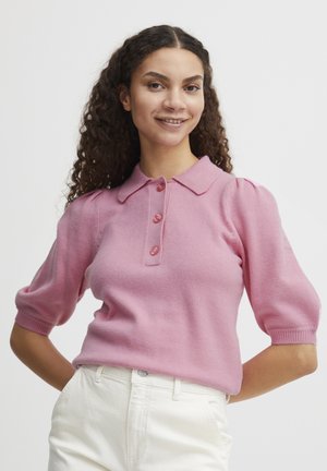 Rosa kurzärmeliger Pullover mit Kragen und drei Knöpfen, mit gepufften Schultern und weicher Textur. Kombiniert mit weißen Hosen.