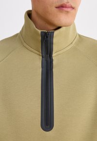 Μακρυμάνικη μπλούζα σε χακί χρώμα με ψηλό γιακά, διαθέτει μαύρο φερμουάρ κατά μήκος του μπροστινού μέρους και υφαντό υλικό με αντίθετες ραφές.