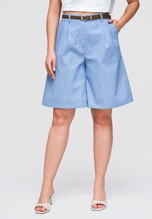 Kvinde iført lyseblå vidde-short med smalle striber, brun bælte, hvid top og beige åbne hælede sandaler.