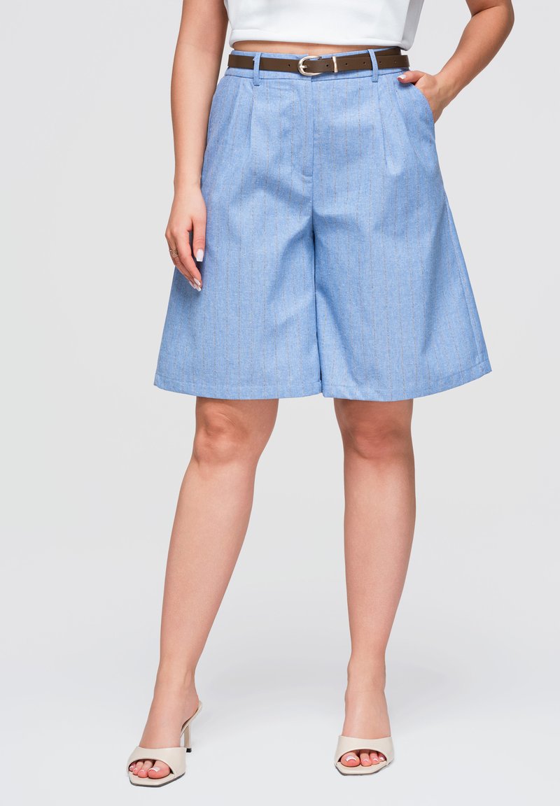 Femme portant un short ample bleu clair à fines rayures, une ceinture marron, un haut blanc et des sandales à talons ouvertes beige.