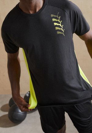 Homme en tenue de sport noire et jaune néon tenant son t-shirt avec deux ballons médicinaux noirs au sol derrière lui.