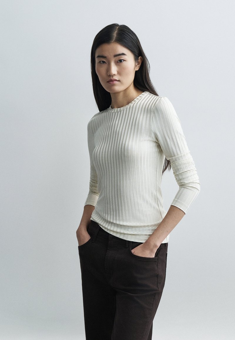 Mlada ženska z dolgimi temnimi lasmi, oblečena v kremno rebrasto dolgimi rokavi in črne hlače, stoji z rokami v žepih pred enobarvnim sivim ozadjem.