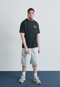 T-shirt en coton noir avec logo, shorts gris clair, chaussettes blanches et baskets grises avec accents. Debout sur un tapis bleu contre un mur clair.