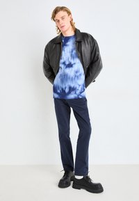 Veste oversize en cuir noir, pull bleu tie-dye, pantalon navy et chaussures noires épaisses avec une semelle épaisse. Modèle posé sur un fond uni.
