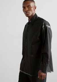 Sort skjorte med lange ærmer og knaplukning, der har en glinsende bånddetalje på skuldrene og en rund afslutning. Glat stoftekstur.