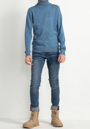 Blauer Rollkragenpullover, taillierte Passform, gerippte Bündchen und Saum. Kombiniert mit schmal geschnittenen Jeans und braunen Schnürstiefeln mit robusten Sohlen.