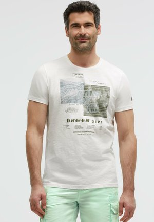 Mann trägt ein weißes T-Shirt mit Grafiken, Texten und Bildern, kombiniert mit hellgrünen Cargoshorts, steht vor einem einfarbigen Hintergrund.