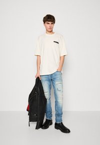 Kremfarget oversized T-skjorte med svart logo, slitte lyseblå jeans og svarte ankelstøvler. Holder en svart skinnjakke.