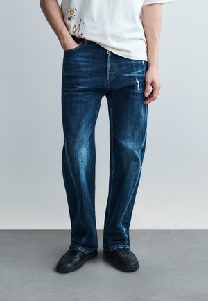 Personne portant un jean bleu droit avec une légère usure, des baskets noires et un t-shirt blanc orné de motifs brodés rouges et jaunes.