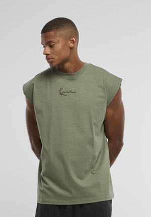 Hombre con camiseta sin mangas verde oliva con pequeño logo de texto en el pecho, mirando hacia la izquierda con las manos detrás de la espalda, fondo liso gris claro.