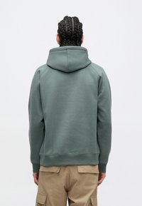 Duks s kapuljačom u prigušenoj zelenoj boji, izrađen od mekanog materijala. Ima kangaroo džep, rebrasti rub i rukave; glatka tekstura s udobnim krojem.