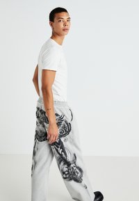 Gray sweatpants with black graphic prints, elastic waistband, side pockets; paired with a plain white t-shirt and black shoes.