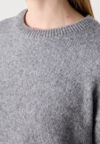 Gros plan sur une personne portant un pull en tricot doux gris clair avec un col rond côtelé et une texture visible.