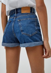 Pantalones cortos de mezclilla de tiro alto, con dobladillos enrollados y dos bolsillos traseros. Color azul claro con una etiqueta de cuero en la cinturilla.