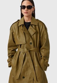 Olijf groen leren trenchcoat met een ceintuur in de taille, dubbelzijdige knopen, grote revers en zijzakken. Bevat flap- en knoopdetails.