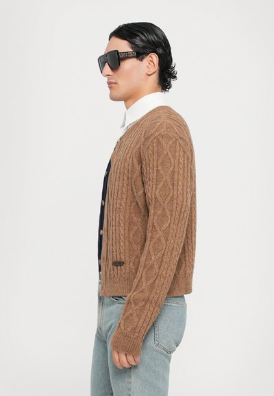 Brun kabelstickad cardigan med ribbade ärmslut, marinblå skjorta under, ljus tvättade denimjeans och överdimensionerade svarta solglasögon.