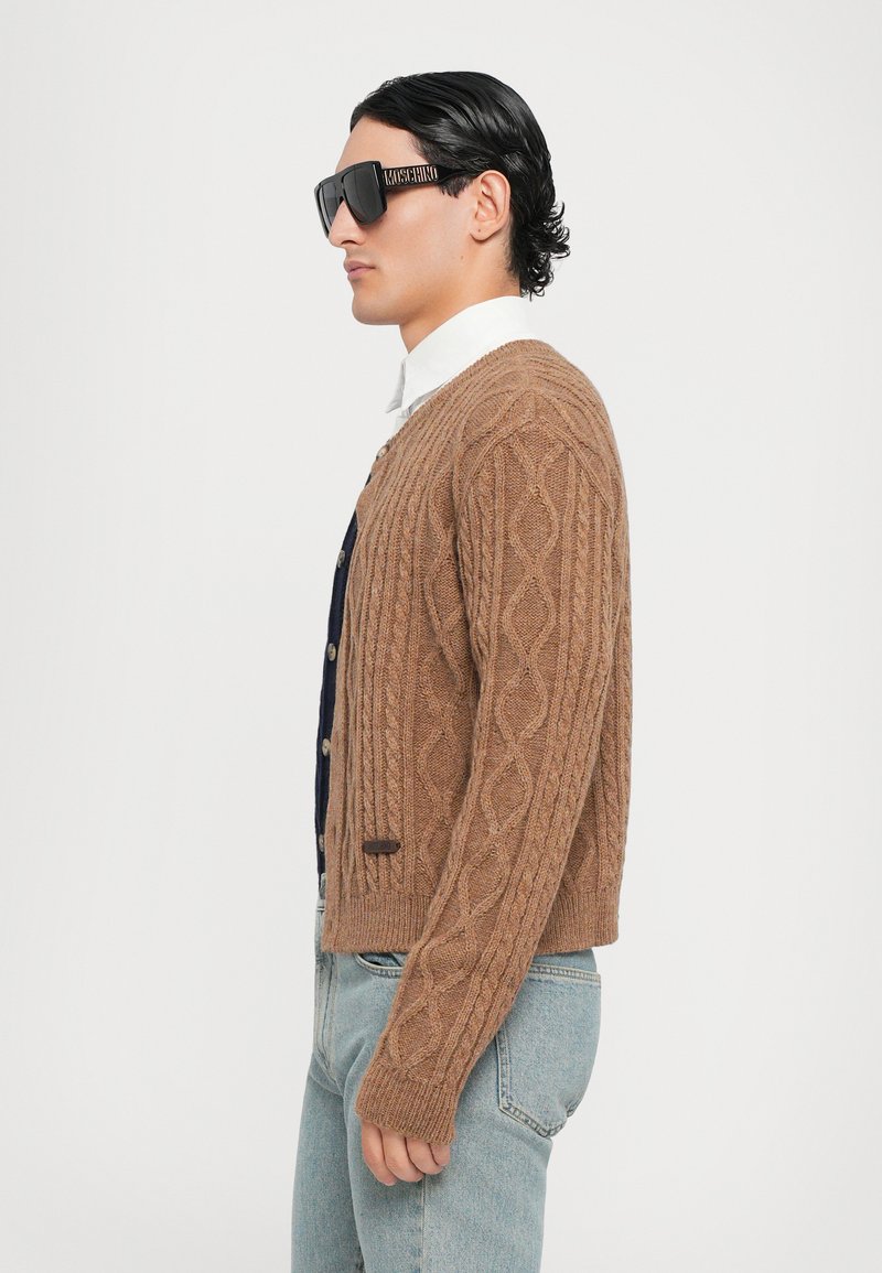 Bruine kabelgebreide cardigan met geribbelde manchet, een marineblauw overhemden eronder, lichtgewassen spijkerbroek en oversized zwarte zonnebril.