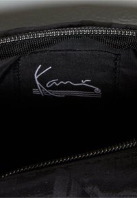 Zwarte stoffen binnenkant van een tas met een zilveren rits en een label met "Kamo" in witte cursieve tekst tegen een donkere achtergrond.