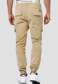 Pantalons cargo beiges avec une taille élastique, poches latérales, jambes fuselées et poignets élastiques, associés à des baskets blanches avec des accents marins.