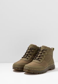 Par de botas altas verde oliva con suelas cosidas en negro y amarillo, empeines de tejido texturizado y cordones marrones sobre una superficie blanca.
